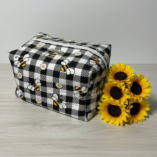 Black and white checkered fabric box with bee pattern next to sunflowers on a wooden surface.